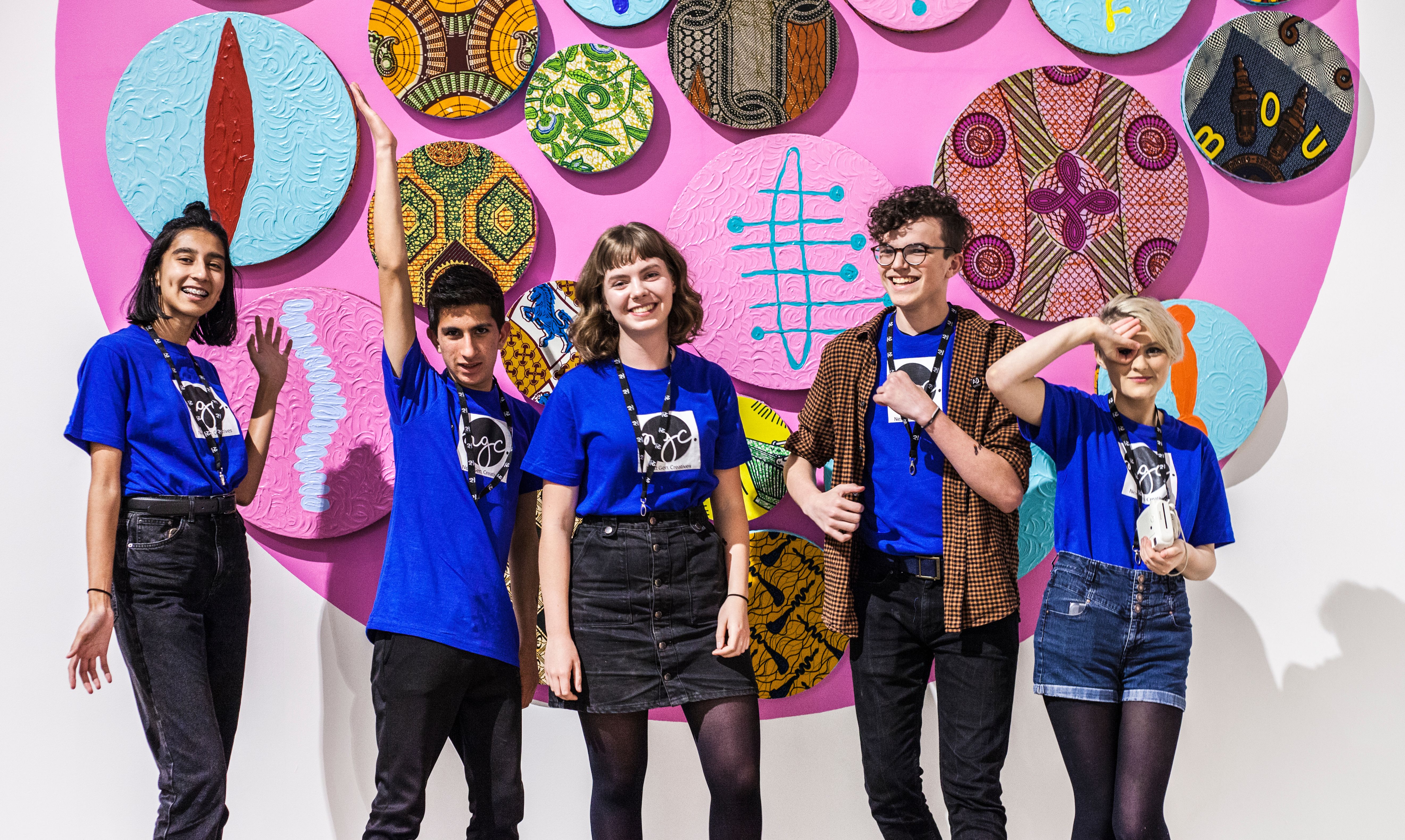 Five young people wearing electric blue tshirts infront of a bright pink background looking happy