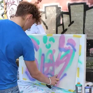 Photo of teenage boy with short hair wearing an electric blue t.shirt bending over spray painting a canvas